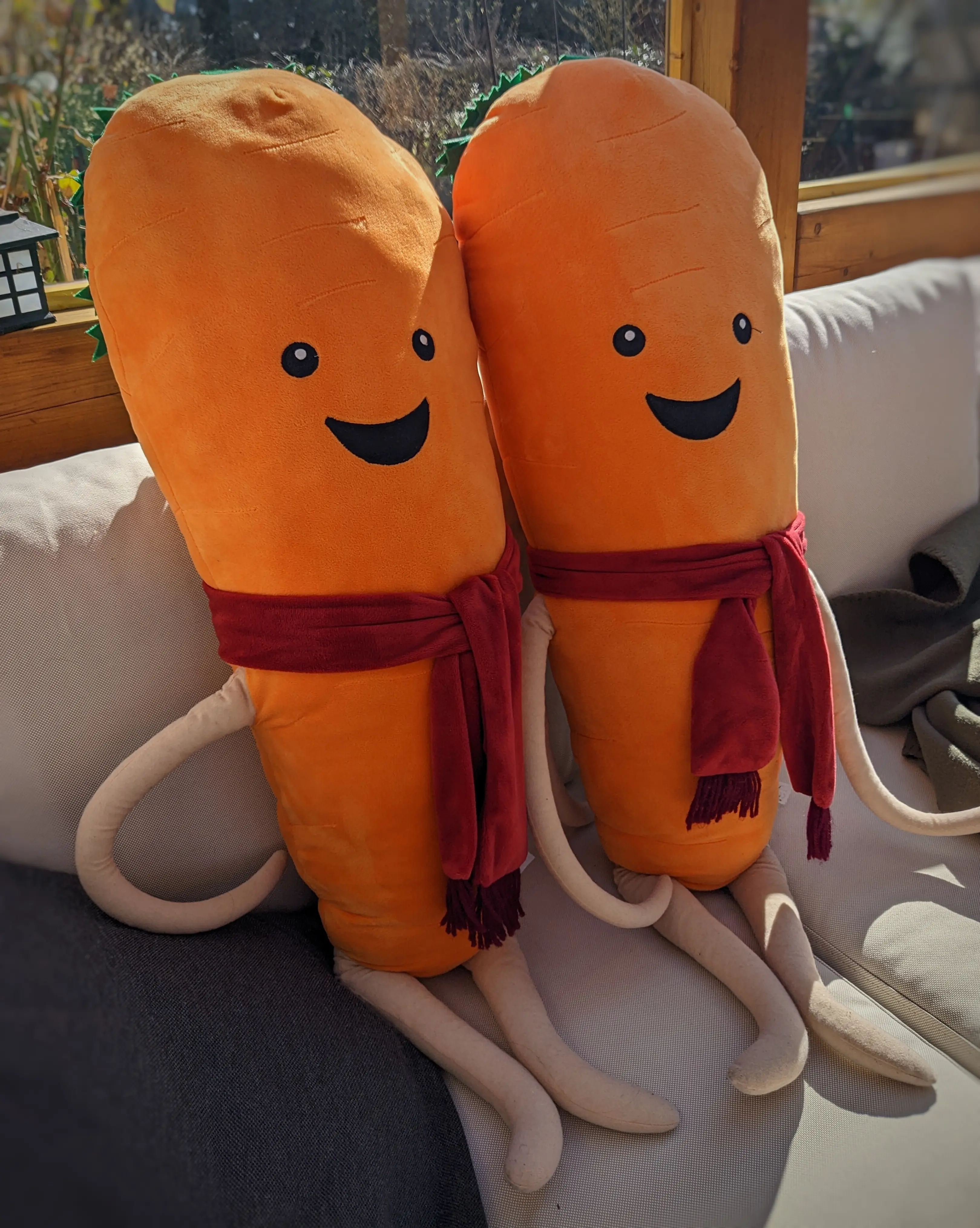Two plush carrots with beige arms and legs sitting on a white sofa.
They have red scarfs wrapped around their "necks" and a smile on their face.
Their carrot greens are hanging off their head on the back, like hair.
