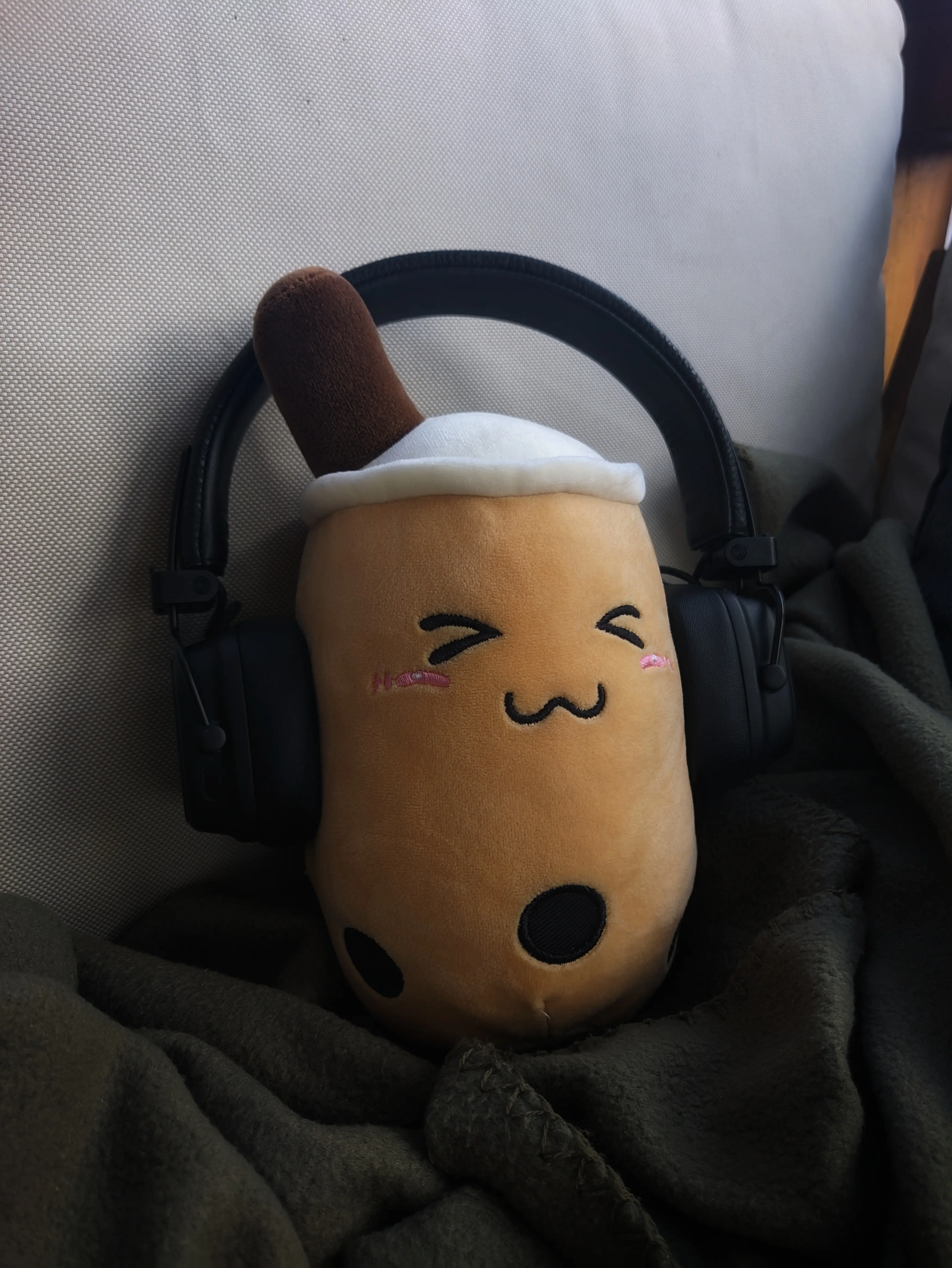 Light-brown plush bubble tea with black boba and a cute stitched on facial expression.
It is sitting on a dark green blanket and wearing a black pair of on-ear headphones.
